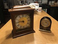 Both clocks displayed side by side on a kitchen table with visible details of size and style.