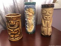 Three ceramic tiki cups: brown glaze tiki, two-tone tiki with carvings, and tall tiki surfer girl cup with chips.