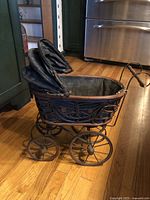 Side view of black Victorian-style doll carriage showing metal frame with intricate detailing, fabric lining, and large metal spoked wheels on wooden floor.