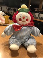 Front view of vintage baby clown plush showing full body in striped blue and white outfit with red yarn hair, red polka-dot bow tie, yellow and green polka dot hat, sitting on a surface.