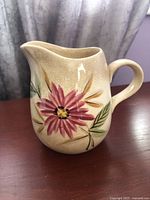 Front angled view of vintage Kasuga Japanese creamer showing pink floral design with green and brown leaves and visible crazing on beige background.
