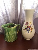 Green glazed pottery pitcher and floral transfer vase together on table by curtain