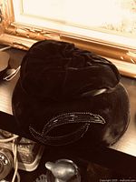 Black velvet Victorian-style hat displayed on a wooden surface beneath a large gold frame, showing the gathered crown and beadwork detail