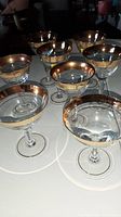Eight clear stemware glasses with broad gold rims arranged on a white surface. Six glasses have tall stems and typical wine glass bowls, two have shorter stems and wider coupe-style bowls.