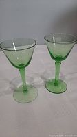 Two green Depression glass liqueur glasses side by side on a white cloth surface against a gray background.