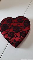 Photo showing a heart shaped gift box with a black lace floral patterned fabric lid over a red base, placed on a white surface.