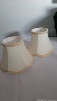 Two small cream-colored lampshades with scalloped and braided trim shown from side angle.