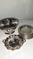 Silverplate Rogers bowl alongside a stack of 13 side dishes and a small Saxon plate, laid out on white cloth.