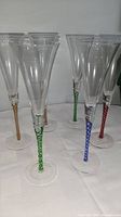 Six clear champagne flutes arranged showing different colored twisted spiral stems (green, blue, red, amber) on a white cloth surface.