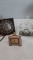 Three clocks displayed on a white cloth surface: a black Westclox electric clock with cord, a clear glass Soos mantel clock with white dial, and a small pink enamel travel clock with gold accents.