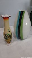 Photo of two art glass vases on a white cloth background. Left vase has ruffled top with marbled colors, right vase is white with vertical colorful stripes.