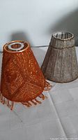 Photo shows two vintage beaded lampshades, orange and grey, sitting on a white textured surface.