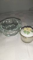 Photo of two vanity jars on a tablecloth; one is a heavy crystal jar with faceted clear glass and the other a smaller ribbed glass jar with a white ceramic lid decorated with yellow roses and green leaves.