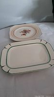 Photo showing both plates on a white cloth, rectangular plate in front with green and black trim, round plate behind with floral design.