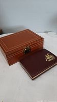 Both vintage jewellery boxes shown on white surface, the larger brown box with detailed stitching and smaller maroon box with gold ship emblem