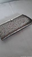 Silver glitter clutch purse lying flat on a light-colored textured fabric surface. Visible is the front side showing a curved edge design with centrally placed snap closure.