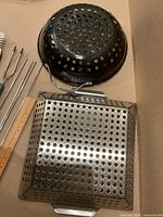 Square perforated stainless steel grilling basket, round black enamel perforated grilling basket and two stainless steel kebab sticks alongside a wooden ruler for size reference.