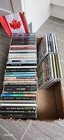 Photo showing a cardboard box with 27 Canadian music CDs stacked in it, along with a small Canadian flag. The CDs include a variety of artists and albums, all in jewel cases.
