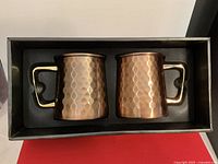 Two copper-tone Moscow Mule mugs with hammered texture and black handles, placed in original black packaging.