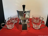 Front view of Bialetti Moka pot with four glass cups with metal handles arranged around it on a red surface.