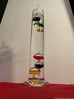 Full view of Galileo thermometer showing clear glass cylinder with colorful floats inside on red background.