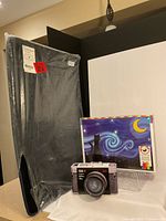 Three items displayed: black Beckos puzzleboard wrapped in plastic, Eurographics Classic Cameras 550-piece puzzle tin shaped like a vintage camera, and 300-piece vintage camera puzzle with night sky design.