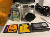 Photo showing Nikon CoolPix 4300 digital camera with lens cap on, two memory cards (Lexar 256 MB and Nikon Lexar), rechargeable Nikon battery, and charger on a tabletop, with camera box in the background.