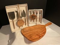 Photo showing olivewood serving board along with two boxed sets of cheese utensils and forks tied with rustic string.