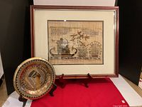 Framed Egyptian papyrus art with boat scene and hieroglyphics, and the embossed metal decorative plate on stands