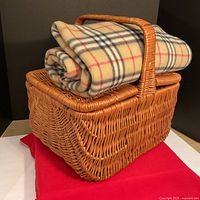 Wicker picnic basket with plaid blanket folded on top seen from angled front.