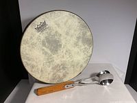Front view of REMO Fiberskyn 3 frame drum propped up vertically with a pair of metal musical spoons with wooden handles laying alongside it.