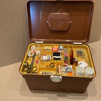 Open brown sewing box with two yellow trays showing assorted sewing threads in different colors, needles, pins, safety pins, thimbles, measuring tape, and other small sewing supplies.