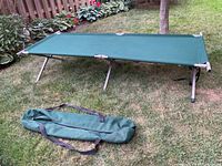 Green outdoor folding camping cot set up on grass next to garden bed, with green carry bag laid out nearby.