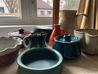 Front angle view of nine pottery pieces on a table in front of a window showing a mix of pitchers, bowls, lidded dish and small vases in multi-colored glazes including teal, brown, orange, and cream.