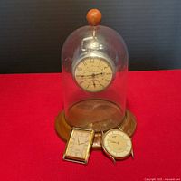 Vintage pocket watch mounted inside a glass dome with a wooden base, with two additional watch heads placed in front, all on a red surface.