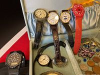 Close up view of four watches with leather and plastic bands and assorted jewelry including coins and costume pieces in the jewelry box.