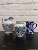 Photo showing the three antique blue and white ceramic pitchers together on a black surface against a white brick background, showcasing size and design variations.