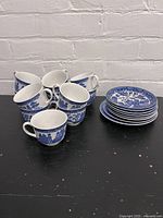Grouped view of 11 Blue Willow cups and 8 matching saucers on black surface against white brick wall.