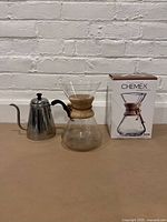Wide shot showing Chemex coffee maker with wood collar and leather tie, stainless steel gooseneck kettle with black handle, and original Chemex box on a beige surface against a white brick background.