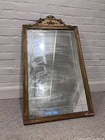 View of the mirror leaned against a white brick wall showing the full frame and decorative top with carved floral and scroll details.