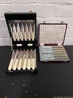 Lot showing two antique cutlery sets, one black box with six forks with white handles and one red box with six butter spreaders with wood-patterned handles.