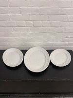 Three white ironstone serving platters displayed side-by-side on black surface against white brick wall, showing size differences and decorative embossed edges