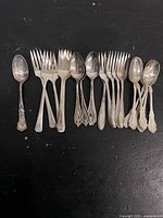 Top view of the silver plated dessert spoons and forks grouped by style on black surface.