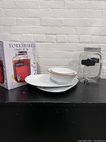 Photo of two ceramic platters with two ceramic bowls and glass drink dispenser with box on left side, against a white brick wall