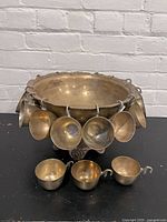 Front view of the brass punch bowl with multiple punch cups hanging on its sides and three cups in front on black surface with a white brick background.