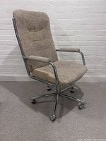 Front and side view showing beige textured fabric upholstery, padded armrests, and chrome metal frame with wheels.