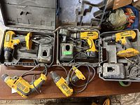 Photo of six DeWalt power tools including cordless drills with batteries, corded drills, an angle grinder, two battery chargers, and three plastic carrying cases on a wooden table.