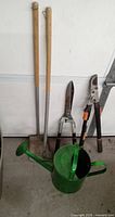 Photo showing two wooden-handled garden hoes, a green metal watering can, a pair of hedge trimmers, a tree pruner, and a pooper scooper against a white wall.