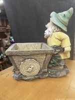 Front-side view of a gnome planter wearing a green hat and yellow tunic pushing a decorative wooden cart planter