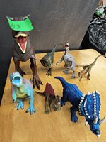 Seven dinosaur figurines arranged on a wooden tabletop, showing variety in size and color.